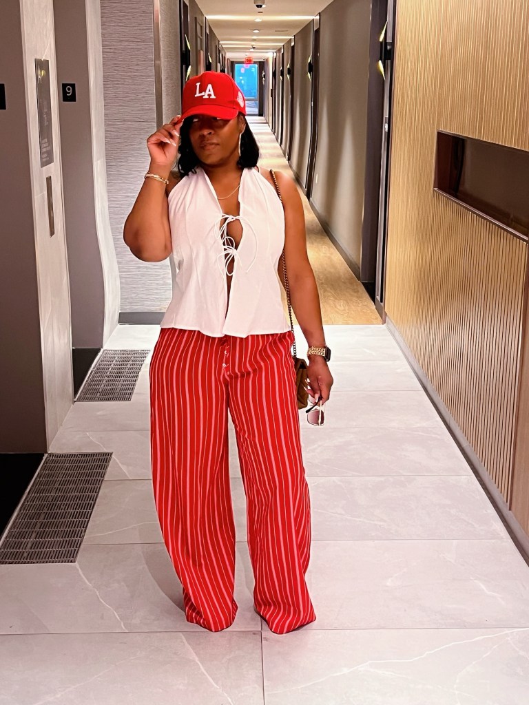 A person stands in a hotel hallway wearing red striped pants and a white open-front top, paired with a red LA cap. The setting features modern decor with soft lighting.